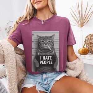 May include: A mauve-colored t-shirt featuring a black and white image of a cat in a mugshot pose, smoking a cigarette and holding a sign that reads "I HATE PEOPLE". The shirt is paired with denim shorts.