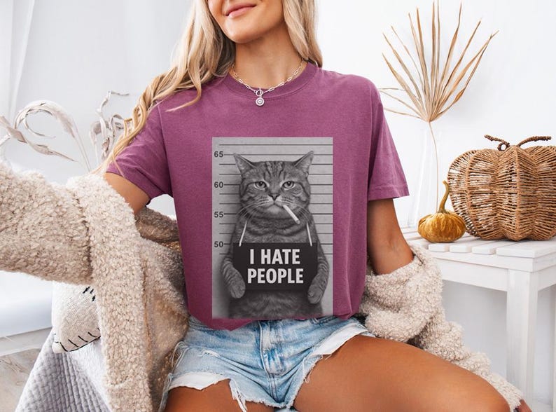 May include: A mauve-colored t-shirt featuring a black and white image of a cat in a mugshot pose, smoking a cigarette and holding a sign that reads "I HATE PEOPLE". The shirt is paired with denim shorts.