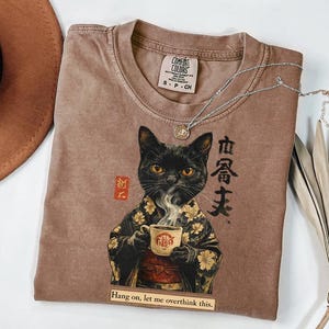 May include: A brown Comfort Colors t-shirt featuring a black cat in a kimono holding a coffee cup. The shirt has Japanese text and the phrase "Hang on, let me overthink this." A silver necklace and a dried plant are also visible.