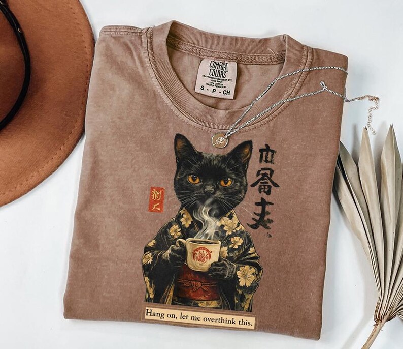 May include: A brown Comfort Colors t-shirt featuring a black cat in a kimono holding a coffee cup. The shirt has Japanese text and the phrase "Hang on, let me overthink this." A silver necklace and a dried plant are also visible.
