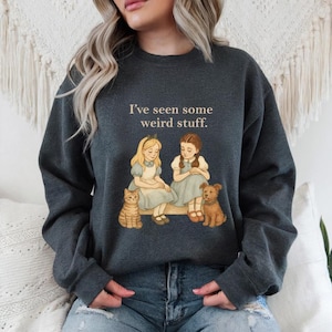 Puede incluir: Sudadera gris oscuro con la frase "I've seen some weird stuff." El gráfico muestra a dos chicas, un gato y un perro. Las chicas visten vestidos y están sentadas en un banco. El gato es naranja y el perro es marrón.