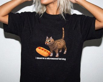 May include: A black short-sleeved t-shirt featuring a graphic of a brown tabby cat standing next to a hot dog in a bun with yellow mustard. Below the cat and hot dog, the white text reads: I deserve a microwaved hot dog.