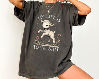 May include: Dark gray t-shirt with the text "MY LIFE IS TOTAL SHIT" and a graphic of a lamb. The lamb is white with a black collar and is surrounded by flowers. The shirt has short sleeves and a relaxed fit.