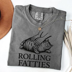 May include: A gray t-shirt with a graphic of a cat being petted, with the words "Rolling Fatties" in black. A gold necklace and a brown hat are also visible. The shirt is made of a soft material.