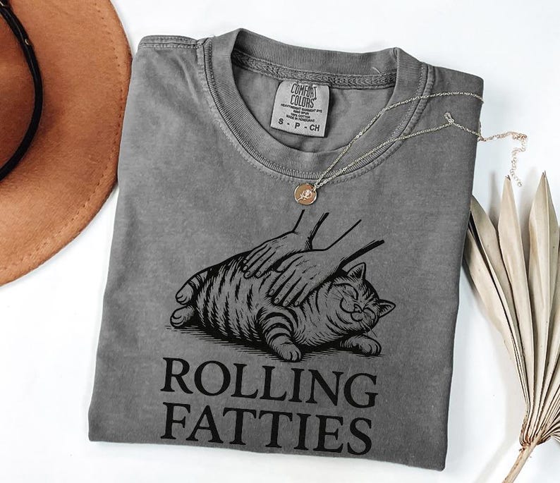 May include: A gray t-shirt with a graphic of a cat being petted, with the words "Rolling Fatties" in black. A gold necklace and a brown hat are also visible. The shirt is made of a soft material.