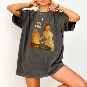 May include: Dark gray t-shirt with the text "I'M FINE" above a vintage illustration of a woman in a dress and apron tending a pan with flames. The graphic is in shades of yellow, orange, and brown.