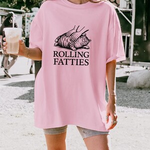 May include: A light pink t-shirt with the text "ROLLING FATTIES" in black. The graphic shows a cat being petted. The shirt is a loose fit.