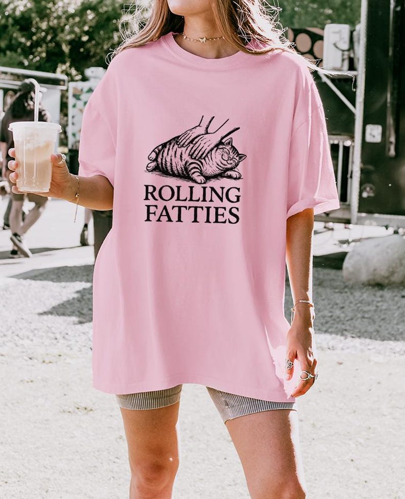 May include: A light pink t-shirt with the text "ROLLING FATTIES" in black. The graphic shows a cat being petted. The shirt is a loose fit.