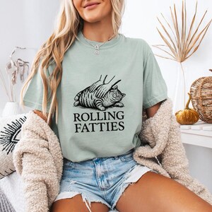 May include: A light green t-shirt with a black graphic of a cat being petted, with the text "ROLLING FATTIES" below. The shirt is worn with denim shorts. The overall theme is casual and humorous.