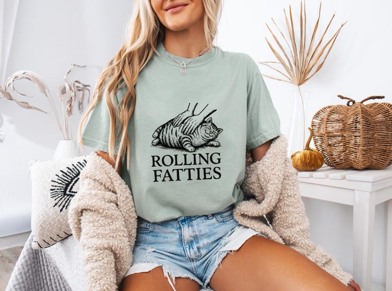 May include: A light green t-shirt with a black graphic of a cat being petted, with the text "ROLLING FATTIES" below. The shirt is worn with denim shorts. The overall theme is casual and humorous.