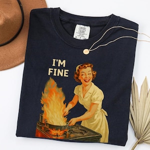 May include: Navy blue t-shirt with the text "I'M FINE" above a retro illustration of a woman cooking with flames. A gold necklace with a small pendant is draped on the shirt. A brown hat is visible in the background.