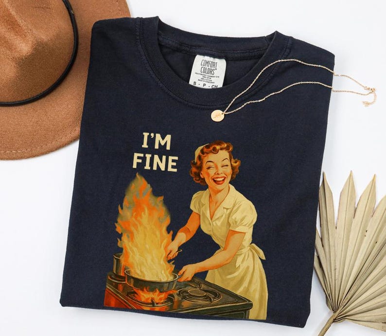 May include: Navy blue t-shirt with the text "I'M FINE" above a retro illustration of a woman cooking with flames. A gold necklace with a small pendant is draped on the shirt. A brown hat is visible in the background.