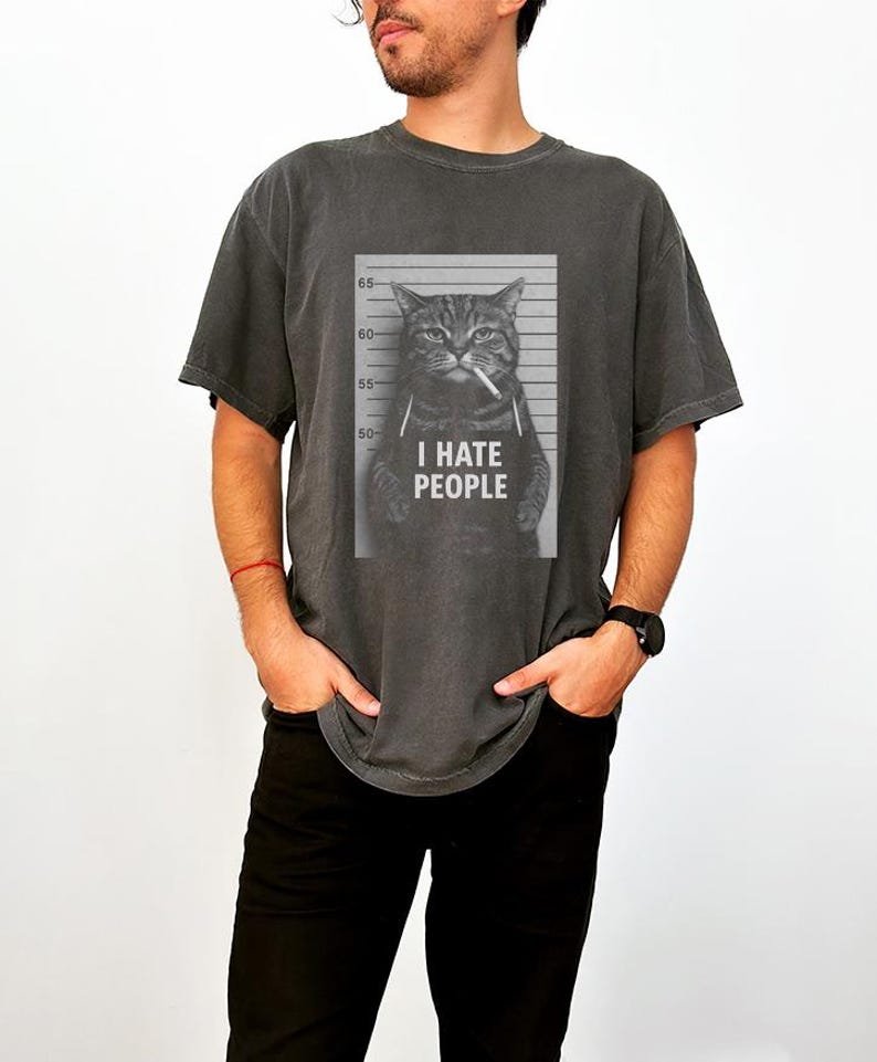 May include: Dark gray t-shirt featuring a black and white image of a cat in a mugshot. The cat is holding a cigarette and the text "I HATE PEOPLE" is below the image. The shirt has short sleeves and a crew neck.