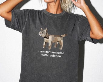 May include: A charcoal gray short-sleeve t-shirt featuring a geometric cat illustration in shades of brown and tan. Below the cat, the text reads, "I am contaminated with radiation" in white, sans-serif lettering. The fabric has a washed, vintage appearance.