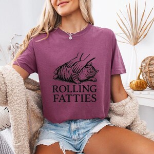 May include: A maroon t-shirt with a graphic of a cat being petted, with the text "ROLLING FATTIES" in black. The shirt is worn with denim shorts and a cream-colored cardigan.