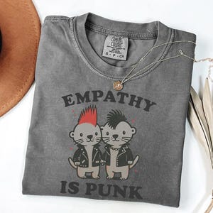 Puede incluir: Una camiseta gris Comfort Colors con el texto "EMPATHY IS PUNK" y un gráfico de dos nutrias de dibujos animados con chaquetas de cuero. Un collar dorado con un colgante está sobre la camiseta. Un sombrero marrón está en la esquina superior izquierda.
