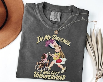 May include: A charcoal gray t-shirt featuring a graphic of a child in a hat and polka dot skirt pushing a toy stroller with a teddy bear inside. The text reads, In My Defense, I Was Left Unsupervised. The shirt has a crew neck and short sleeves.