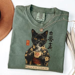 May include: Sage green t-shirt featuring a black cat illustration in a kimono holding a cup of coffee. The shirt has the text "Hang on, let me overthink this." and "COMFORT COLORS" printed on the tag.