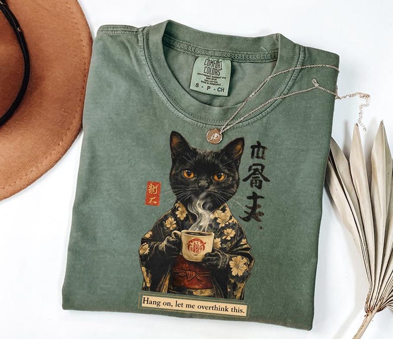 May include: Sage green t-shirt featuring a black cat illustration in a kimono holding a cup of coffee. The shirt has the text "Hang on, let me overthink this." and "COMFORT COLORS" printed on the tag.