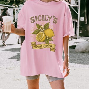 May include: A light pink t-shirt with the text "Sicily's Finest Lemons" in a vintage style. The graphic features lemons, leaves, and flowers. The shirt is a relaxed fit.