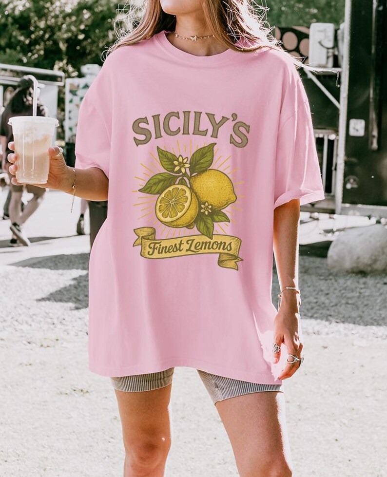 May include: A light pink t-shirt with the text "Sicily's Finest Lemons" in a vintage style. The graphic features lemons, leaves, and flowers. The shirt is a relaxed fit.