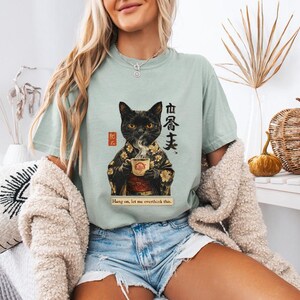 May include: A light green t-shirt featuring a black cat in a kimono holding a teacup. The shirt has Japanese text and the phrase "Hang on, let me overthink this." The cat design is detailed with gold accents.