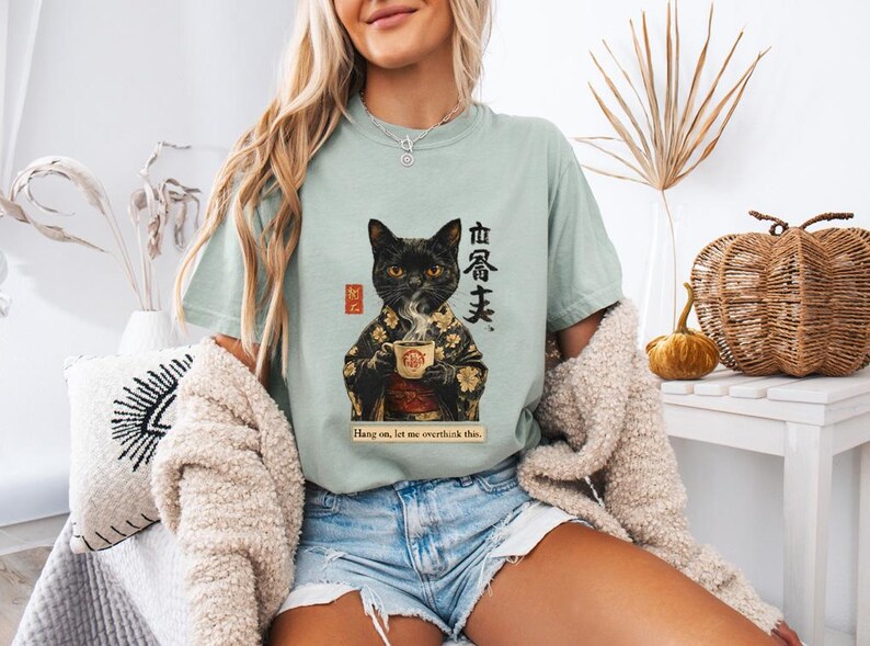 May include: A light green t-shirt featuring a black cat in a kimono holding a teacup. The shirt has Japanese text and the phrase "Hang on, let me overthink this." The cat design is detailed with gold accents.