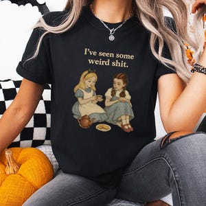 May include: Black t-shirt with the phrase "I've seen some weird shit." The shirt features an illustration of two girls, one in a blue dress and the other in a blue dress, seated with a teapot and a plate of food. The shirt is worn by a person.