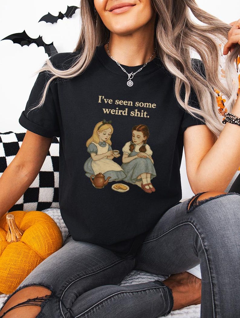 May include: Black t-shirt with the phrase "I've seen some weird shit." The shirt features an illustration of two girls, one in a blue dress and the other in a blue dress, seated with a teapot and a plate of food. The shirt is worn by a person.