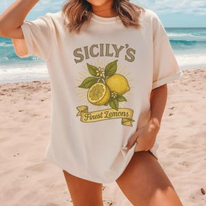 May include: Cream-colored t-shirt featuring a lemon graphic with the text "Sicily's Finest Lemons." The design includes a whole lemon, a sliced lemon, leaves, and flowers. The shirt has short sleeves and a relaxed fit.