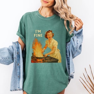 May include: Teal-colored t-shirt with the text "I'M FINE" above a vintage illustration of a woman smiling while cooking with flames. The shirt is a relaxed fit and has short sleeves.