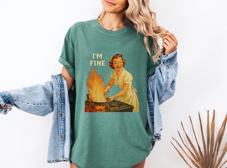 May include: Teal-colored t-shirt with the text "I'M FINE" above a vintage illustration of a woman smiling while cooking with flames. The shirt is a relaxed fit and has short sleeves.