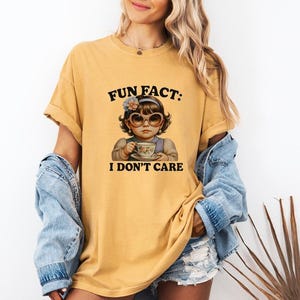 Könnte beinhalten: Senfgelbes T-Shirt mit einer Grafik eines Mädchens mit Sonnenbrille, das eine Teetasse hält. Der Text auf dem Shirt lautet "FUN FACT: I DON'T CARE". Das T-Shirt hat kurze Ärmel und eine lockere Passform.