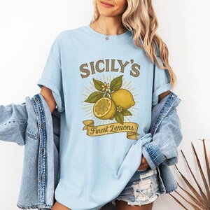 May include: Light blue t-shirt with the text "SICILY'S Finest Lemons" in a vintage style. The graphic features lemons, leaves, and flowers. The shirt is a soft, comfortable material.