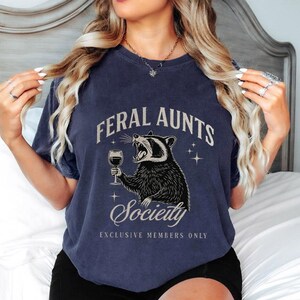 May include: Navy blue t-shirt with the text "FERAL AUNTS Society EXCLUSIVE MEMBERS ONLY" and a graphic of a raccoon holding a wine glass. The raccoon is black and white, and the text is in a vintage style font.