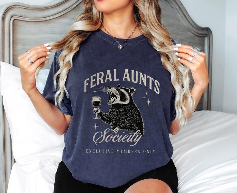 May include: Navy blue t-shirt with the text "FERAL AUNTS Society EXCLUSIVE MEMBERS ONLY" and a graphic of a raccoon holding a wine glass. The raccoon is black and white, and the text is in a vintage style font.