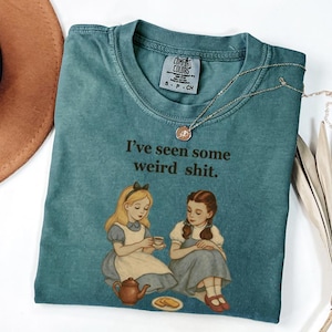 May include: A teal Comfort Colors t-shirt with the phrase "I've seen some weird shit" printed on it. The graphic features illustrations of Alice and Dorothy from Alice in Wonderland and The Wizard of Oz. A gold necklace is draped across the shirt.