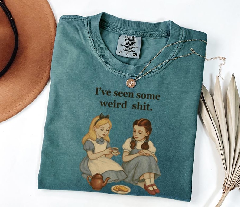 May include: A teal Comfort Colors t-shirt with the phrase "I've seen some weird shit" printed on it. The graphic features illustrations of Alice and Dorothy from Alice in Wonderland and The Wizard of Oz. A gold necklace is draped across the shirt.
