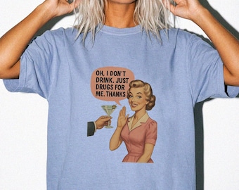 May include: A light blue short-sleeve t-shirt featuring a graphic print of a person in a pink dress declining a martini offered by a hand. A speech bubble above the figure contains the text: OH, I DON'T DRINK. JUST DRUGS FOR ME. THANKS.