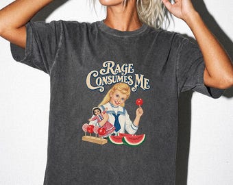 May include: A charcoal grey short-sleeve t-shirt featuring a graphic print of a blonde girl in a sailor-style top holding a doll and a red candy apple. The text "RAGE CONSUMES ME" is printed above, with red lollipops and watermelon slices below.
