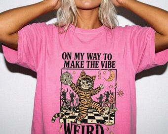 May include: A bright pink t-shirt featuring a graphic of a cat in a tiger-striped onesie holding a disco ball. The text reads ON MY WAY TO MAKE THE VIBE WEIRD. The design includes silhouettes of dancing figures, a crescent moon, and a lava lamp.