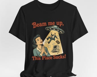 May include: A black t-shirt featuring a graphic print of a person looking up at a UFO abducting a business person. The text reads "Beam me up, This Place Sucks!" in a retro font. The design includes a computer, papers, and a briefcase.