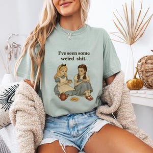 May include: A light green t-shirt with the text "I've seen some weird shit." features an illustration of two girls from Alice in Wonderland. The girls are seated, one holding a teacup. The shirt is paired with denim shorts.
