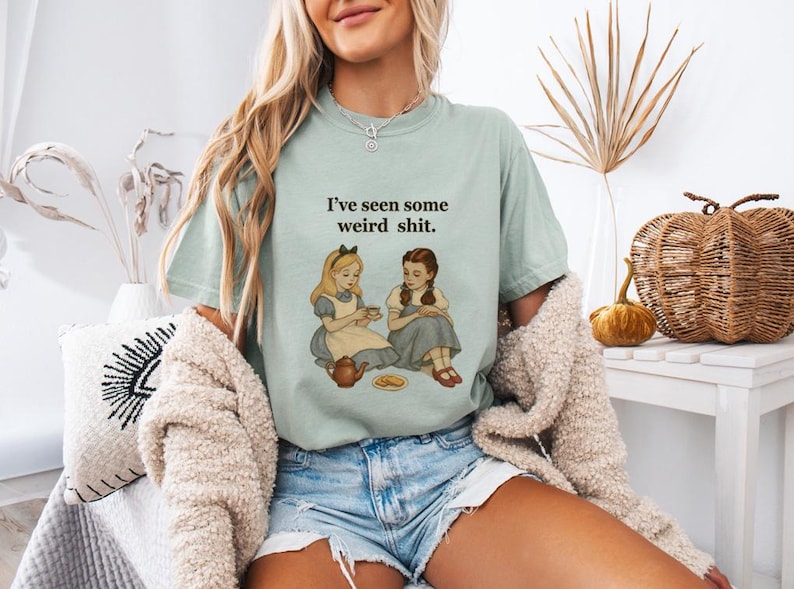 May include: A light green t-shirt with the text "I've seen some weird shit." features an illustration of two girls from Alice in Wonderland. The girls are seated, one holding a teacup. The shirt is paired with denim shorts.