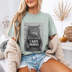 May include: A light green t-shirt featuring a black and white image of a cat in a mugshot, smoking a cigarette, with the text "I HATE PEOPLE" on a sign. The shirt is worn with denim shorts.