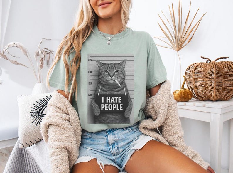 May include: A light green t-shirt featuring a black and white image of a cat in a mugshot, smoking a cigarette, with the text "I HATE PEOPLE" on a sign. The shirt is worn with denim shorts.