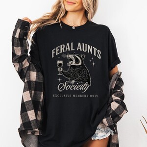 May include: Black t-shirt with the text "FERAL AUNTS Society" and a raccoon graphic holding a wine glass. The raccoon is roaring. The shirt also has the text "EXCLUSIVE MEMBERS ONLY". The person is wearing a black and brown plaid shirt.