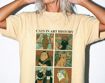 May include: A pale yellow t-shirt featuring a graphic print titled CATS IN ART HISTORY. The design consists of six rectangular panels depicting cats reimagined in famous paintings, including The Scream, Mona Lisa, and Girl with a Pearl Earring.