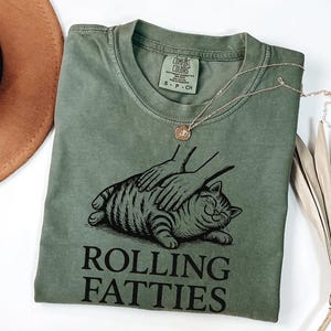 May include: An olive green t-shirt with a graphic of a cat being petted, with the words "ROLLING FATTIES" in black. A gold necklace with a pendant is draped over the shirt. The shirt is folded, and a brown hat is partially visible in the background.