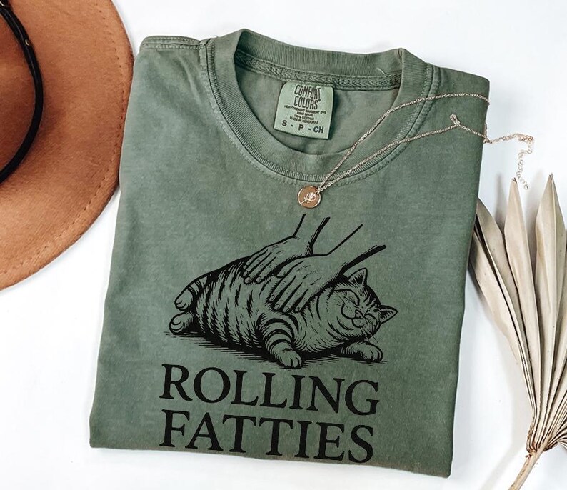May include: An olive green t-shirt with a graphic of a cat being petted, with the words "ROLLING FATTIES" in black. A gold necklace with a pendant is draped over the shirt. The shirt is folded, and a brown hat is partially visible in the background.